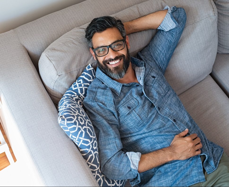 Happy mature man with eyewear and beard relaxing on sofa at home. Handsome latin man with hands behind head sitting on couch in living room. High angle view of cheerful indian man in casual lying on gray couch and looking at camera.