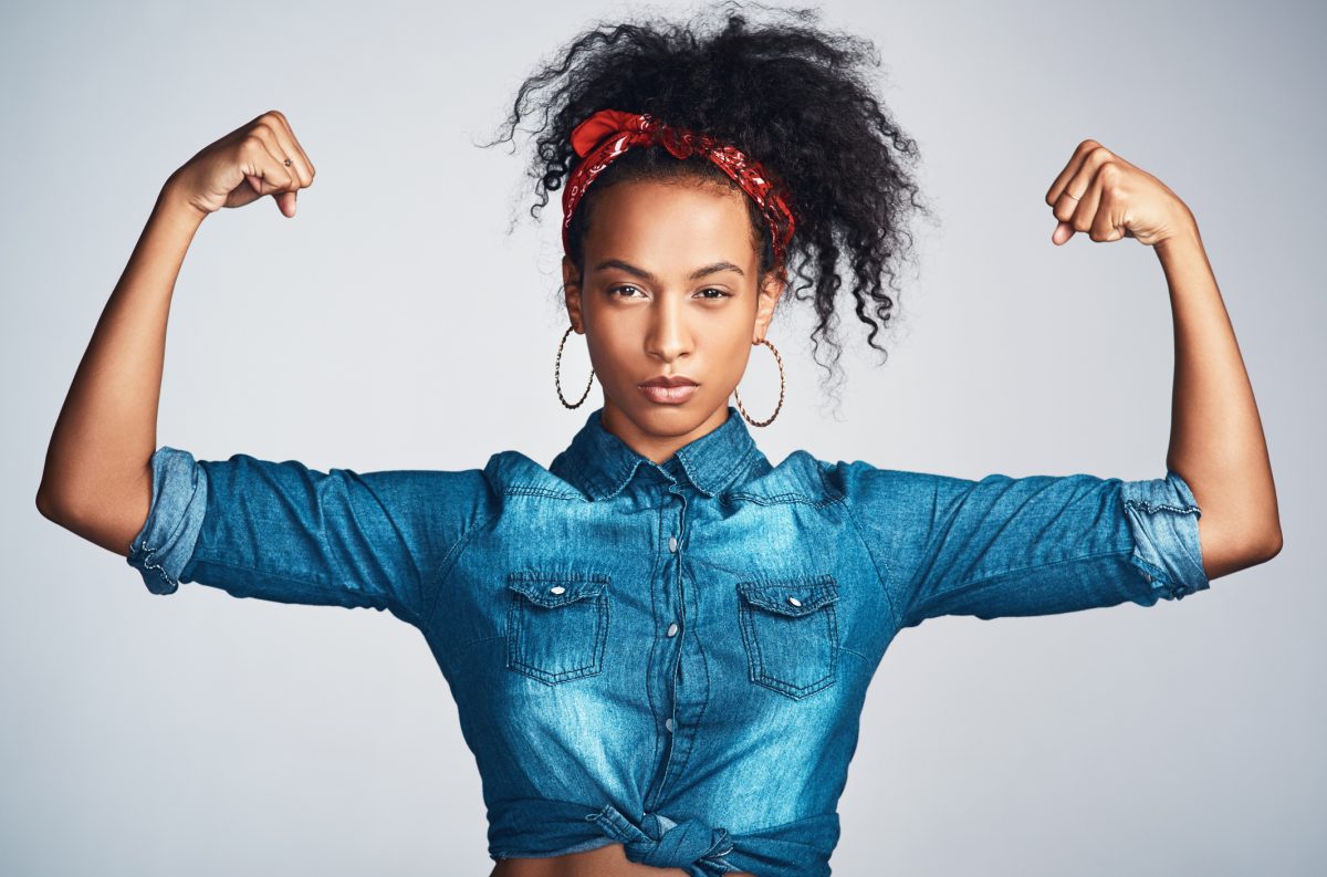 Portrait of a confident young woman wearing denim clothes and looking strong while flexing her biceps against a grey background