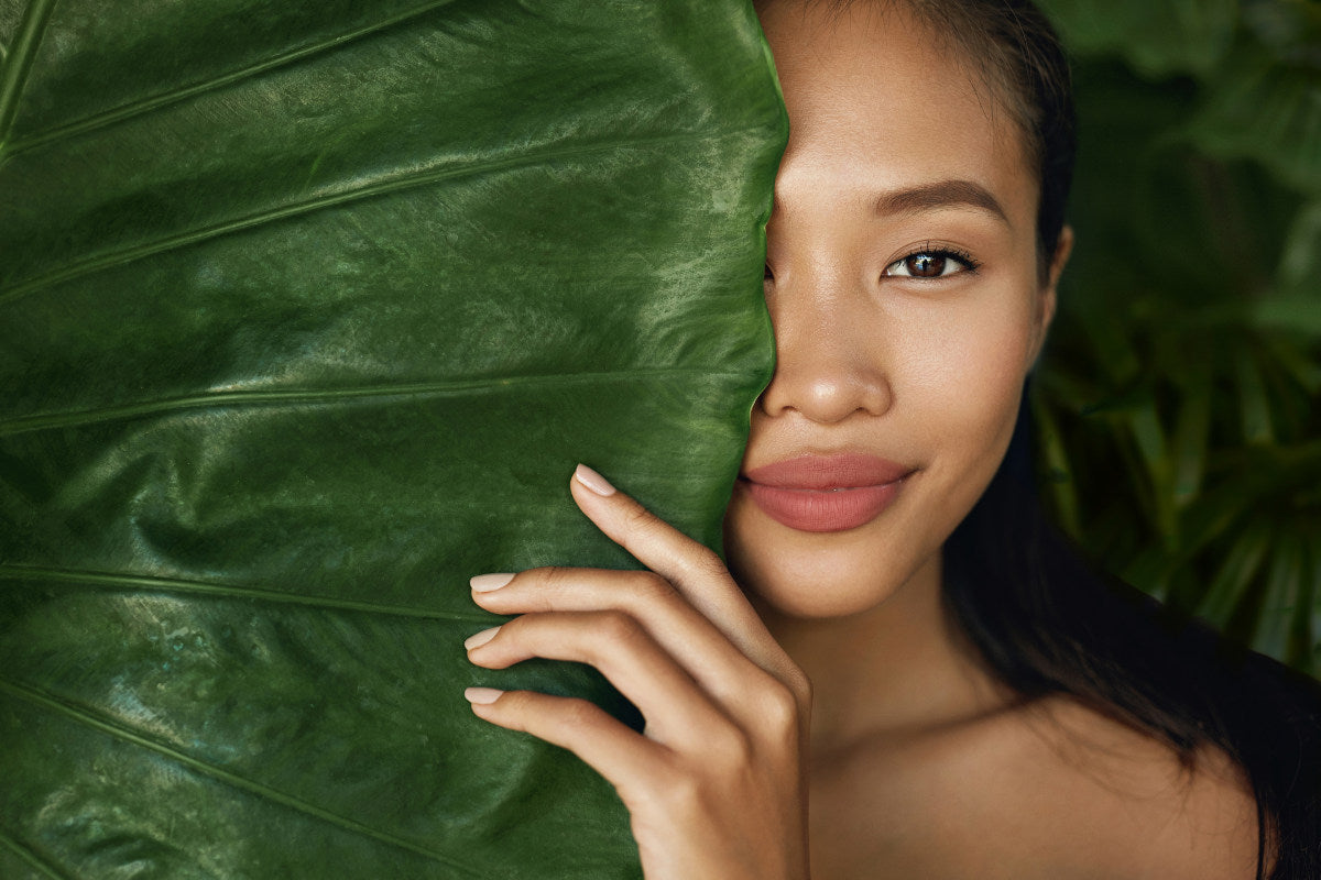 Beauty face. Woman model with natural makeup and healthy skin behind green leaf plant. Portrait of beautiful asian girl with nude nails, big lips and sexy smile in tropical nature