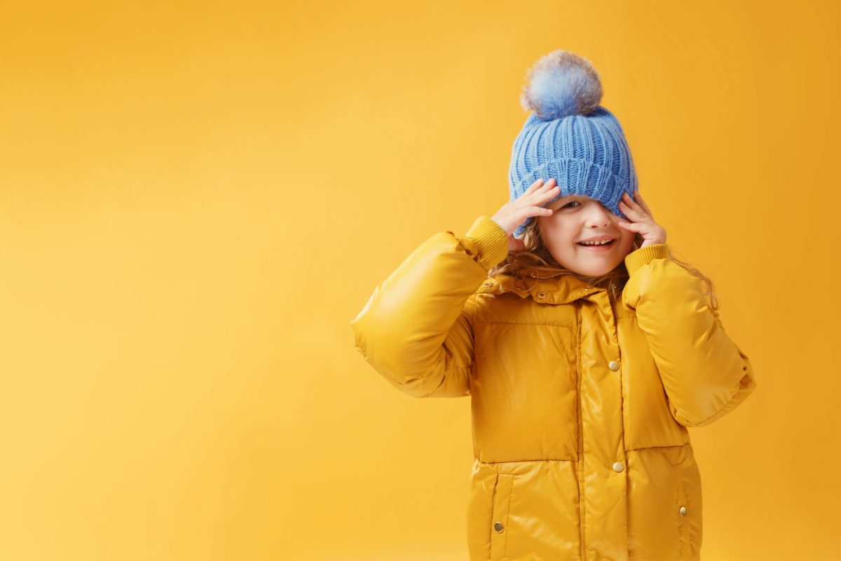 Portrait happy little girl preschooler in a winter jacket and blue hat. Yellow on a yellow background.