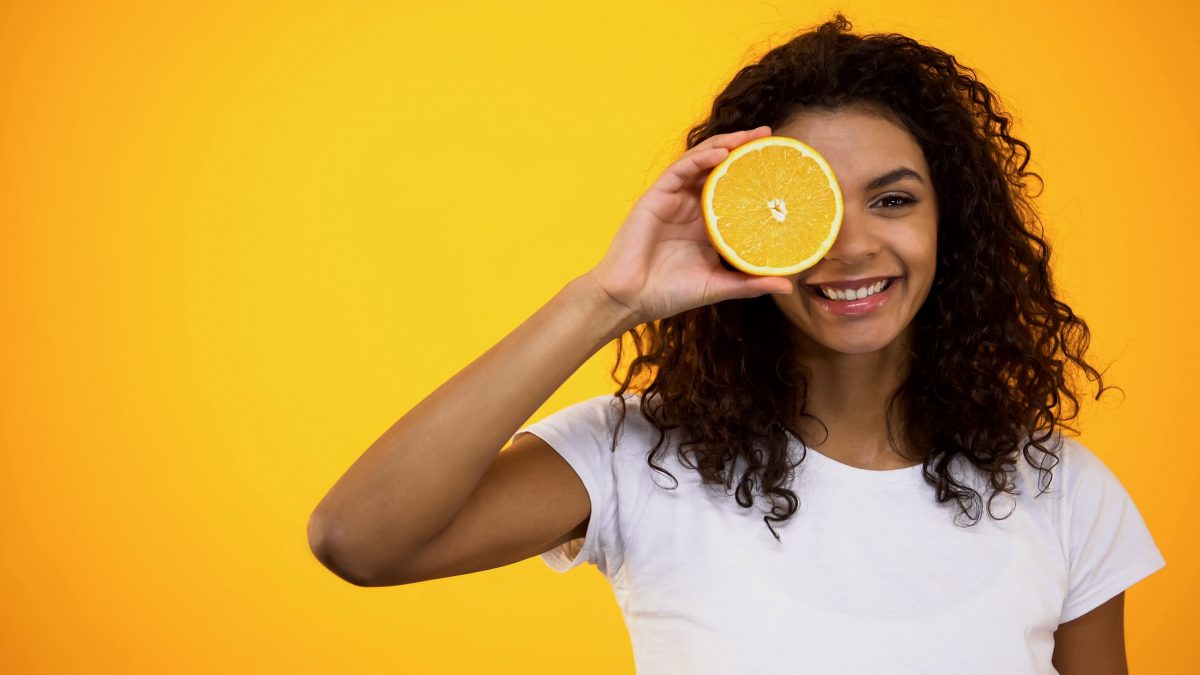 Healthy american woman holding orange half front of eye, vitamins and wellness